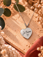 Collier cœur argenté personnalisé avec gravure « Je t’aime » sur pendentif, présenté sur une pierre beige avec fleurs séchées – Atelier Dénésis.